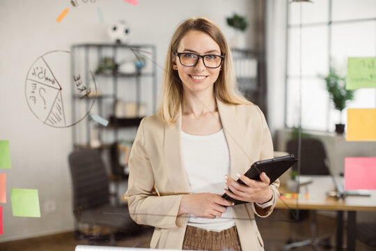 Portrait Of Beautiful Young Woman Standing In Front Of Glass Board And Smiling On Camera. Female Office Worker In Glasses And Stylish Suit Using Digital Tablet In Company.