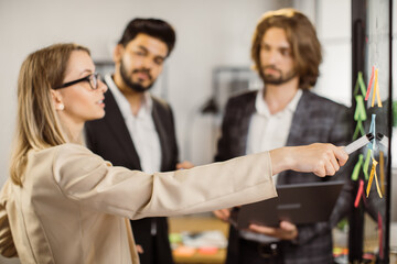 Multicultural team of managers planning and discussing new project in meeting room. Focus on caucasian womans hand pointing with marker on transparent board with diverse notes.