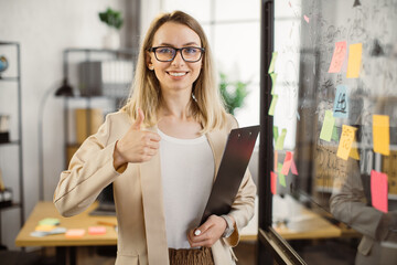 Smiling business lady with clipboard standing near office glass wall with sticky notes and looking...