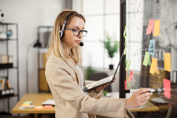 Pensive project manager in headphones drawing strategy plan on glass board during video call on modern laptop. Caucasian woman leading working meeting online at bright office room.
