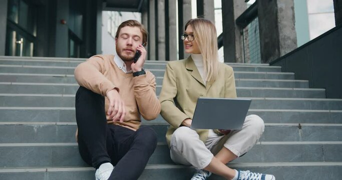Attractive man and woman sitting on stairs in urban city center working on laptop, talking on mobile phone smiling, stylish freelance people using technology outside, making notes. Outside