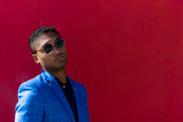 A young man in a blue suit posing with sunglasses on a red wall