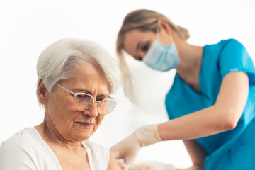 Elderly worried lady with grey hair sitting and having an injection in her arm done by a female nurse in a blue medical uniform. High quality photo