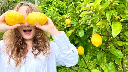 a cheerful teenage girl in a white blouse plays with lemons against the background of a lemon tree she shouts in delight and rejoices. High quality photo