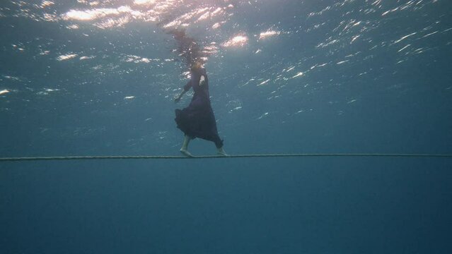 beautiful woman in flowing dress walk rope underwater dark background, she swims to surface of water, dancing in pristine blue ocean water, amazing snorkeling adventure. Armature freediver girl diving