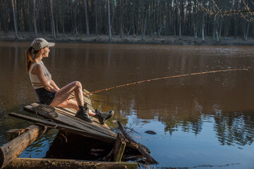 A girl with a fishing rod on a wooden pier is fishing in a forest lake.