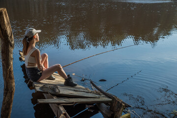 A girl with a fishing rod on a wooden pier is fishing in a forest lake.