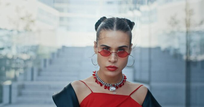 Slow motion portrait of seductive stylish woman wearing red eyeglasses and blouse looking into camera while standing by glass skyscrapers wall. Charming cool fashion female in city street downtown