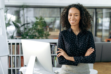 Lovely successful african american curly businesswoman, marketer, lawyer stands near desktop in...