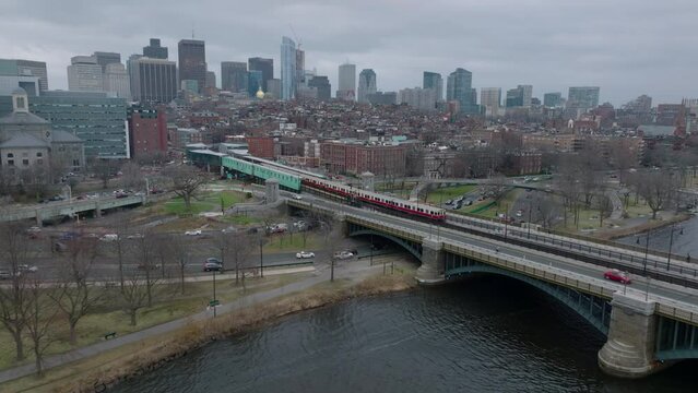 Aerial footage of subway unit crossing river over bridge and stopping in station. Cityscape on cloudy day in background. Boston, USA