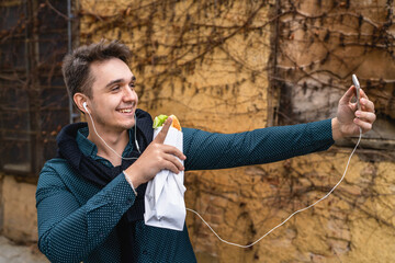 One man standing outdoor in autumn spring or winter day holding sandwich tourist wearing shirt eating while wait outdoor in front of old wall use mobile phone real people copy space fast food concept