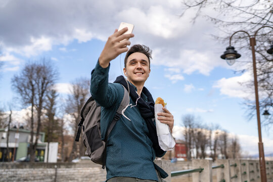 One man standing outdoor in autumn spring or winter day holding sandwich tourist wearing shirt eating on feet stand outdoor in city or town use mobile phone real people copy space fast food concept