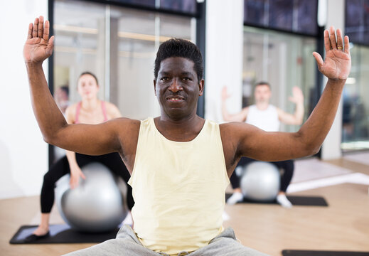 Positive adult African American man maintaining active lifestyle exercising with fitball during group pilates class in fitness studio