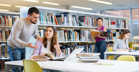 Portrait of two young adult students studying at library using books and laptop, working on common project