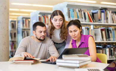 Three adult students work on a laptop and read books in a public library