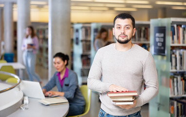 Portrait of a confident student guy, standing in the university library, holding textbooks in his hands - obrazy, fototapety, plakaty