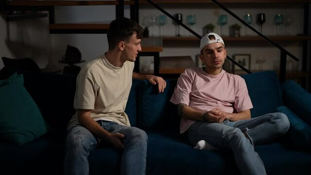 Young Caucasian gay couple arguing in living room at home in darkness. Dissatisfied displeased LGBTQ men talking gesturing yelling indoors. Relationship problems concept