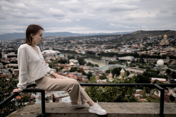 Fototapeta na wymiar Stylish woman in a white sweater and beige pants poses against the backdrop of the city and mountains.