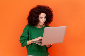 Fototapeta na wymiar Portrait of shocked woman with Afro hairstyle wearing green casual style sweater working on portable computer, looking at display with open mouth. Indoor studio shot isolated on orange background.