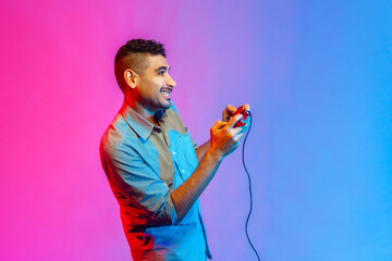 Side view of man in shirt holding in hands red gamepad joystick, grimacing playing video games with...