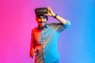 Portrait of smiling man in shirt wearing virtual reality goggles, raising VR headset and screaming with positive expression. Indoor studio shot isolated on colorful neon light background.