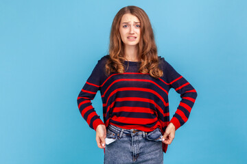 I'm bankrupt! Unhappy poor woman, turning empty pockets inside out and looking frustrated by overspend, lack of money, wearing striped casual sweater. Indoor studio shot isolated on blue background.