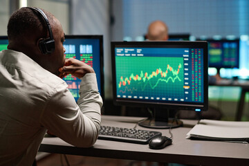 Rear view of tired African American broker sitting in front of computer monitors watching stock trading stats in evening