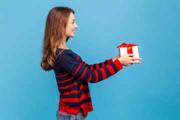 Side view portrait of smiling woman wearing in casual sweater, giving present box, greeting close person with holiday, expressing positive emotions. Indoor studio shot isolated on blue background.