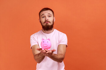 Portrait of sad frustrated bearded man holding piggy bank in hands, being unhappy, has no savings, expressing sorrow, wearing pink T-shirt. Indoor studio shot isolated on orange background.