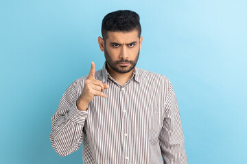 Portrait of serious strict bearded businessman standing with admonishing gesture, scolding for mistake and warning, wearing striped shirt. Indoor studio shot isolated on blue background. - obrazy, fototapety, plakaty
