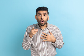 Shocked businessman pointing fingers at camera and looking with big eyes and open mouth, unbelievable news, astonishment, wearing striped shirt. Indoor studio shot isolated on blue background.