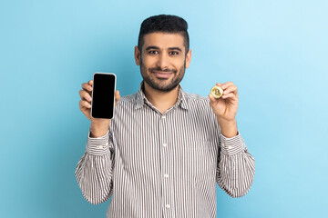 Obraz na płótnie Canvas Bitcoin cryptocurrency payments. Smiling businessman with beard holding btc golden coin and cellphone looks at camera with smile, wearing striped shirt. Indoor studio shot isolated on blue background.