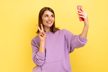 Portrait of optimistic happy woman waving hand to smartphone and taking selfie, showing v sign while making video call, wearing purple hoodie. Indoor studio shot isolated on yellow background.