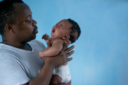 African father holding and comforting sad baby daughter against blue background, holding crying little newborn baby in his arms