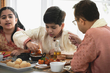 Indian father and daughter and son eating Indian food, using hand, panipuri, curry butter chicken, chicken Tikka, biryani, and naan bread on wooden table, Indian family