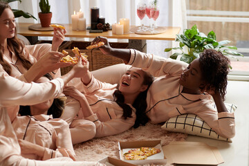 Group of young women toasting with pizza slices when lying on floor in pajamas