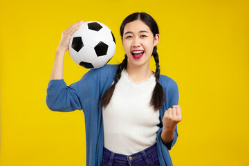 Happy Asian woman football fan cheer up support favorite team with soccer ball isolated on yellow background. People emotions sport family leisure concept.