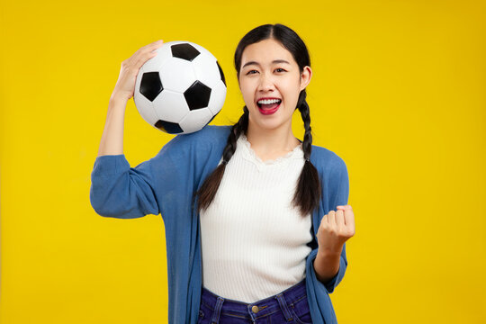 Happy Asian Woman Football Fan Cheer Up Support Favorite Team With Soccer Ball Isolated On Yellow Background. People Emotions Sport Family Leisure Concept.