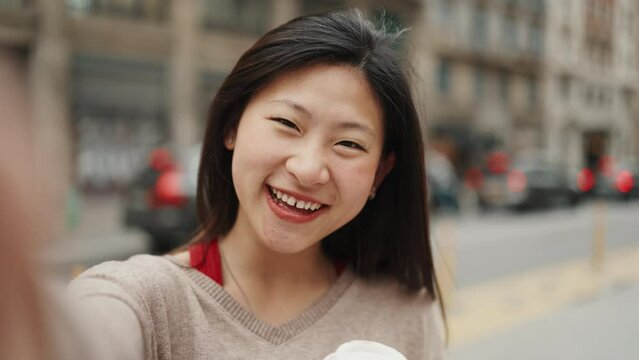 Pretty Asian girl taking self portrait while walking in the city. Long haired smiling woman enjoying a day out in downtown making selfie on phone