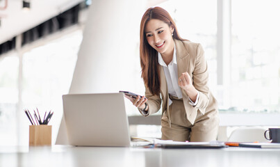 Happy excited successful Asian businesswoman triumphing with a laptop computer smartphone in the workplace office