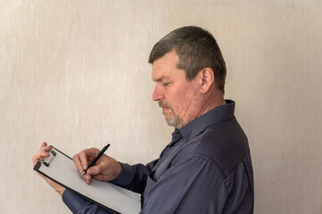 Portrait of a grown man taking notes into a clipboard. Man with short haircut and graying hair. White blank piece of paper clutched in clipboard. Side view. Indoors.