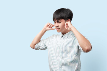 Portrait of young handsome Asian man covering ears with fingers standing blue background.