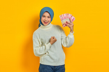 Cheerful beautiful asian woman in white sweater and hijab showing indonesian rupiah banknotes, celebrating luck isolated over yellow background. People islam religious concept