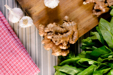 Whole raw fresh ginger place on a wooden chopping board surrounded by kitchen towel, whole garlic and herbs