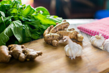 Whole raw fresh ginger place on a wooden chopping board surrounded by kitchen towel, whole garlic and herbs