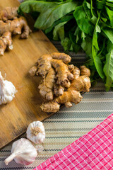 Whole raw fresh ginger place on a wooden chopping board surrounded by kitchen towel, whole garlic and herbs