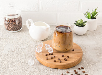 cold dalgona coffee with ice cubes in a large glass on a wooden round board and a cement gray...