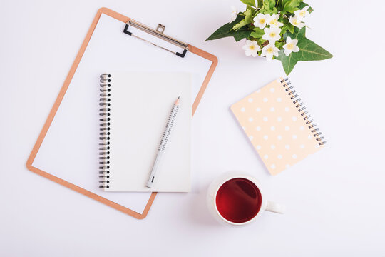 White desktop with clipboard, cup of tea, pencil, notebook and bouquet of spring flowers. Flat lay, top view, mockup