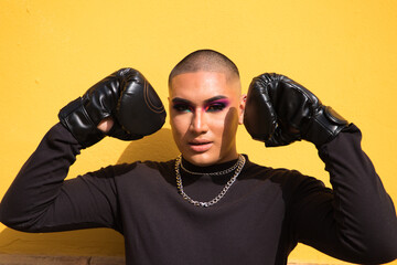 Young non-binary person wears boxing gloves on a mustard yellow background, the person is make up...