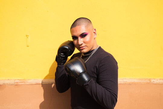 Young non-binary person wears boxing gloves on a mustard yellow background, the person is make up and does different boxing poses. Concept equality, homosexuality, gay, lesbian, gay pride.